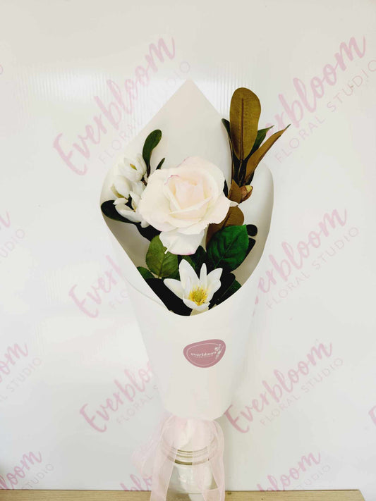 A faux rose with green magnolia leaves arranged in a bouquet, wrapped in a white paper with a ribbon.