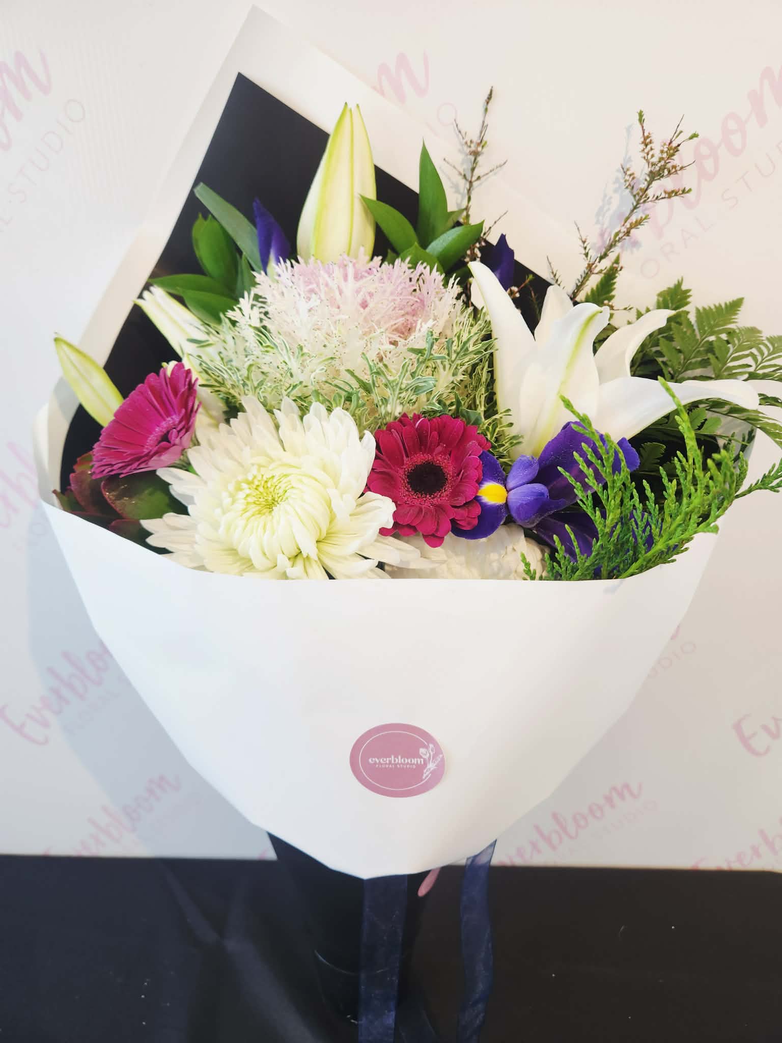 A fresh flower bouquet wrapped in white paper with a pink circular logo, featuring white lilies, pink and white daisies, purple flowers, and green foliage, Everbloom Floral Studio