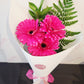 A bouquet of three bright pink gerbera daisies with green fern-like foliage, wrapped in white paper with a pink ribbon and a small card, photographed on a light surface.
Everbloom Floral Studio 