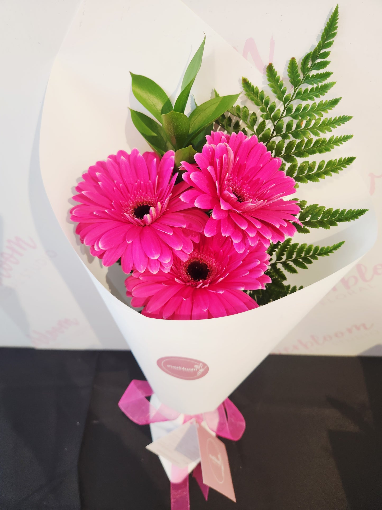 A bouquet of three bright pink gerbera daisies with green fern-like foliage, wrapped in white paper with a pink ribbon and a small card, photographed on a light surface.
Everbloom Floral Studio 
